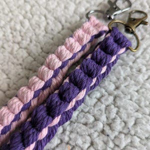 Puede incluir: Dos llaveros de macram&eacute;, uno rosa y otro morado, con cierres plateados y bronce. Los llaveros est&aacute;n hechos de cord&oacute;n de algod&oacute;n trenzado, mostrando un dise&ntilde;o texturizado. El fondo es una superficie suave, blanca y texturizada.