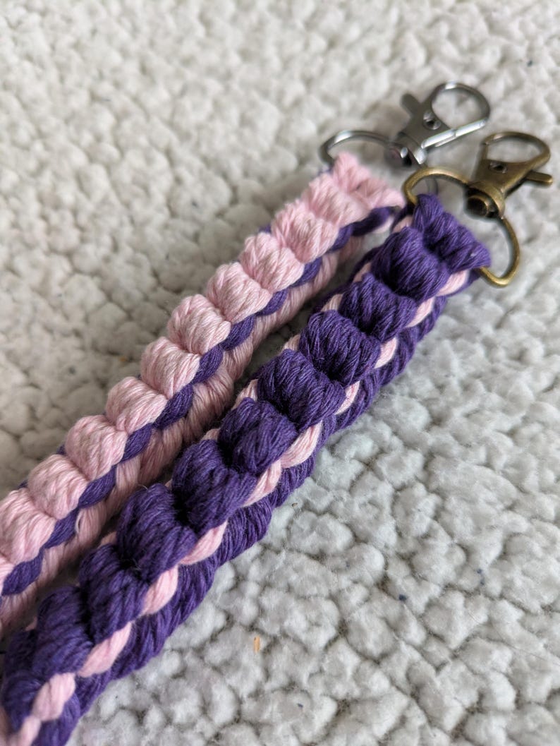 Puede incluir: Dos llaveros de macram&eacute;, uno rosa y otro morado, con cierres plateados y bronce. Los llaveros est&aacute;n hechos de cord&oacute;n de algod&oacute;n trenzado, mostrando un dise&ntilde;o texturizado. El fondo es una superficie suave, blanca y texturizada.