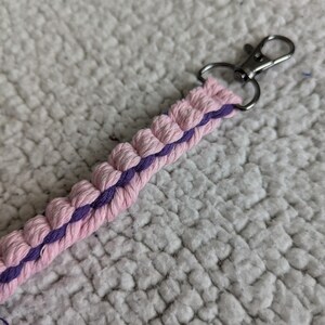Puede incluir: Un llavero de macram&eacute; hecho a mano en rosa y morado. El llavero presenta un dise&ntilde;o trenzado con un cierre plateado. El cord&oacute;n de macram&eacute; rosa est&aacute; acentuado con un centro morado. El fondo es una superficie blanca texturizada.
