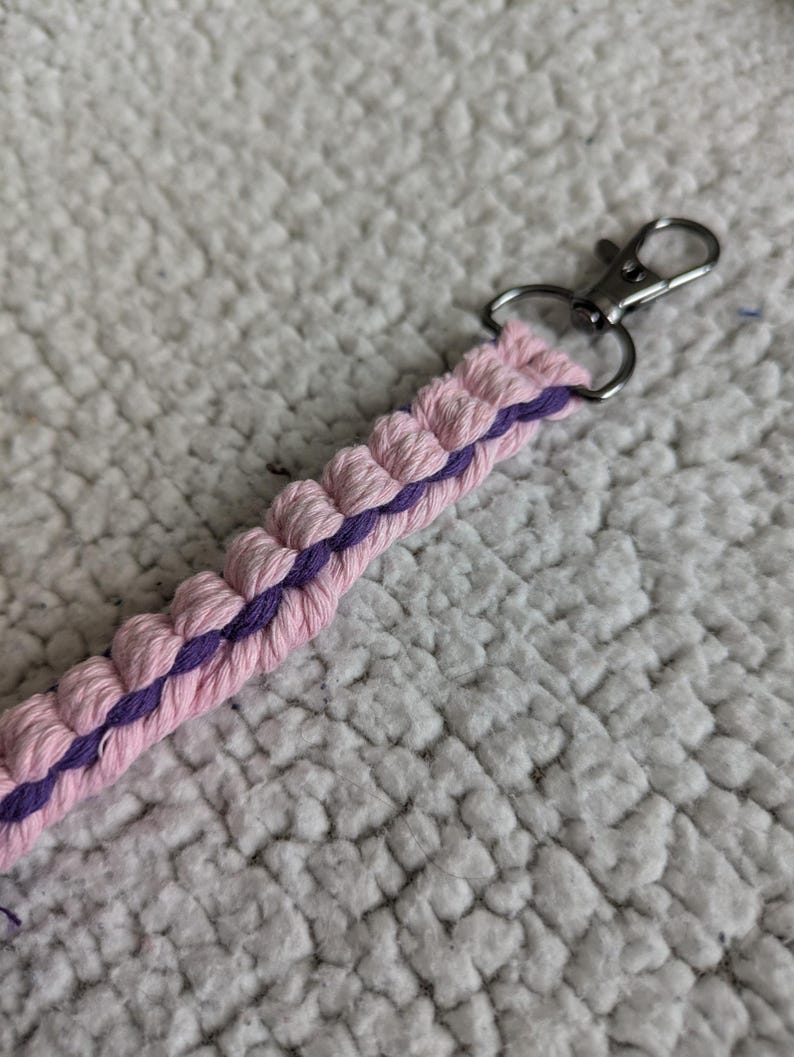 Puede incluir: Un llavero de macram&eacute; hecho a mano en rosa y morado. El llavero presenta un dise&ntilde;o trenzado con un cierre plateado. El cord&oacute;n de macram&eacute; rosa est&aacute; acentuado con un centro morado. El fondo es una superficie blanca texturizada.