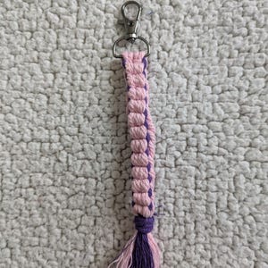 Puede incluir: Llavero de macram&eacute; hecho a mano con dise&ntilde;o rosa y morado. El llavero tiene un cierre plateado y una borla en la parte inferior. El macram&eacute; est&aacute; hecho de cord&oacute;n tejido, con una combinaci&oacute;n de colores rosa y morado.