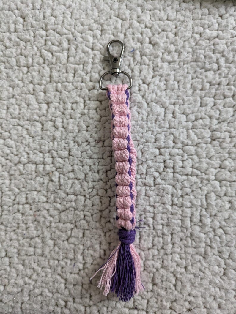 Puede incluir: Llavero de macram&eacute; hecho a mano con dise&ntilde;o rosa y morado. El llavero tiene un cierre plateado y una borla en la parte inferior. El macram&eacute; est&aacute; hecho de cord&oacute;n tejido, con una combinaci&oacute;n de colores rosa y morado.