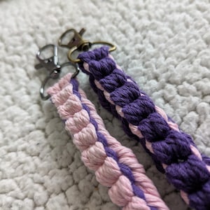 Puede incluir: Dos llaveros de macram&eacute;. Uno es morado y el otro rosa, ambos con cierres plateados y bronce. Los llaveros est&aacute;n hechos de cord&oacute;n de algod&oacute;n trenzado, con un aspecto texturizado. El fondo es una superficie suave y blanca.