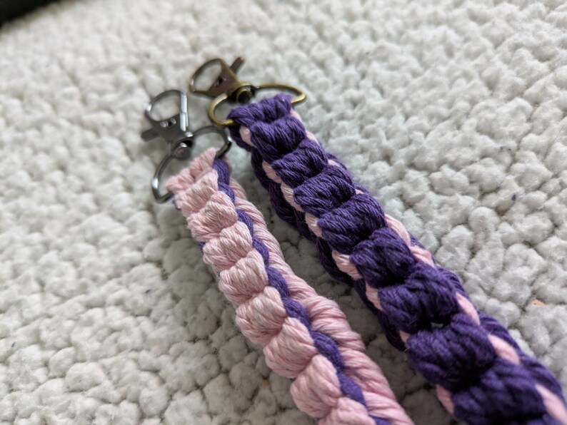 Puede incluir: Dos llaveros de macram&eacute;. Uno es morado y el otro rosa, ambos con cierres plateados y bronce. Los llaveros est&aacute;n hechos de cord&oacute;n de algod&oacute;n trenzado, con un aspecto texturizado. El fondo es una superficie suave y blanca.