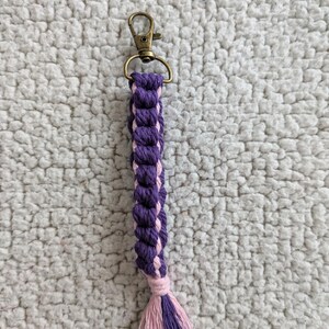 Puede incluir: Un llavero de macram&eacute; hecho a mano con un cierre de mosquet&oacute;n de color bronce. El llavero presenta un dise&ntilde;o trenzado en tonos morados y rosas, con una borla a juego en el extremo. El dise&ntilde;o general es simple y elegante.