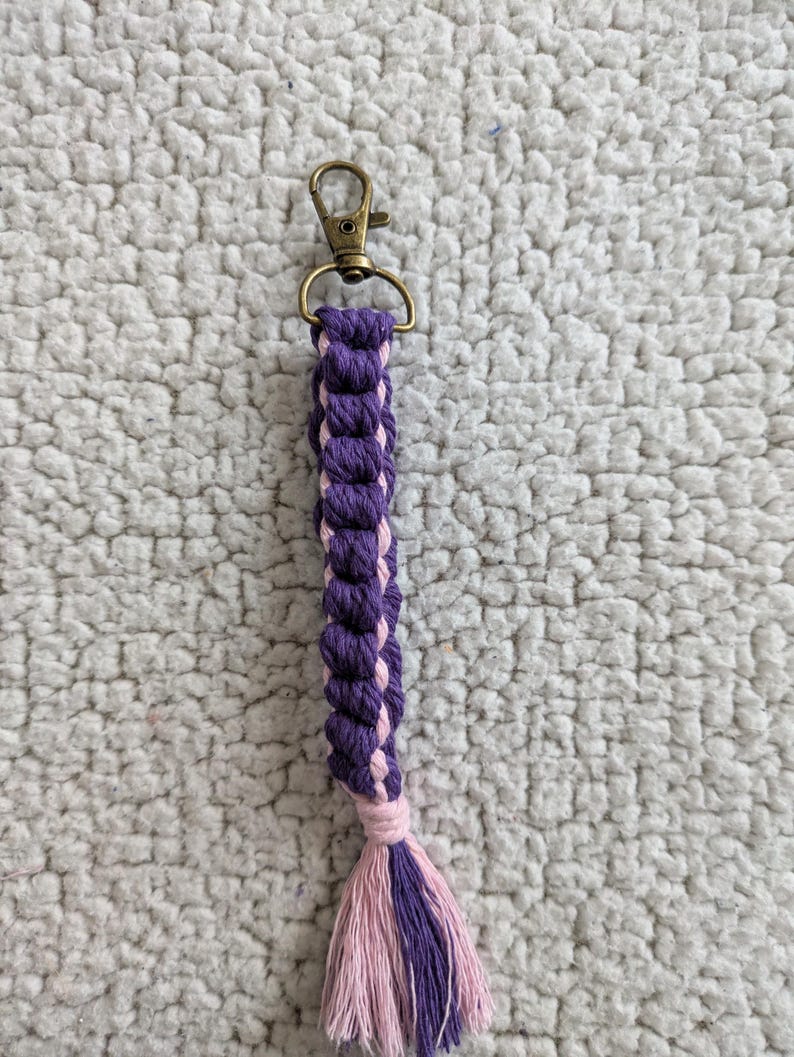 Puede incluir: Un llavero de macram&eacute; hecho a mano con un cierre de mosquet&oacute;n de color bronce. El llavero presenta un dise&ntilde;o trenzado en tonos morados y rosas, con una borla a juego en el extremo. El dise&ntilde;o general es simple y elegante.