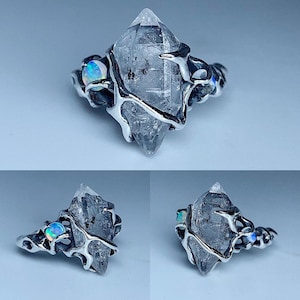May include: A silver ring featuring a large, clear quartz crystal as the centerpiece. The crystal is wrapped in a unique, organic-looking silver setting, with small opal accents on either side. The ring has a modern, artistic design.