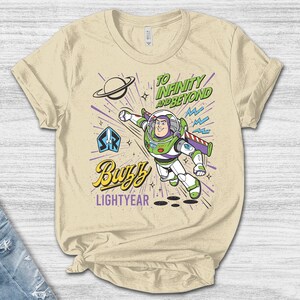 May include: A cream-colored t-shirt featuring a graphic of Buzz Lightyear in a flying pose. The design includes the text "TO INFINITY AND BEYOND" in green, "Buzz" in yellow script, and "LIGHTYEAR" in purple, along with a planet and star icons.
