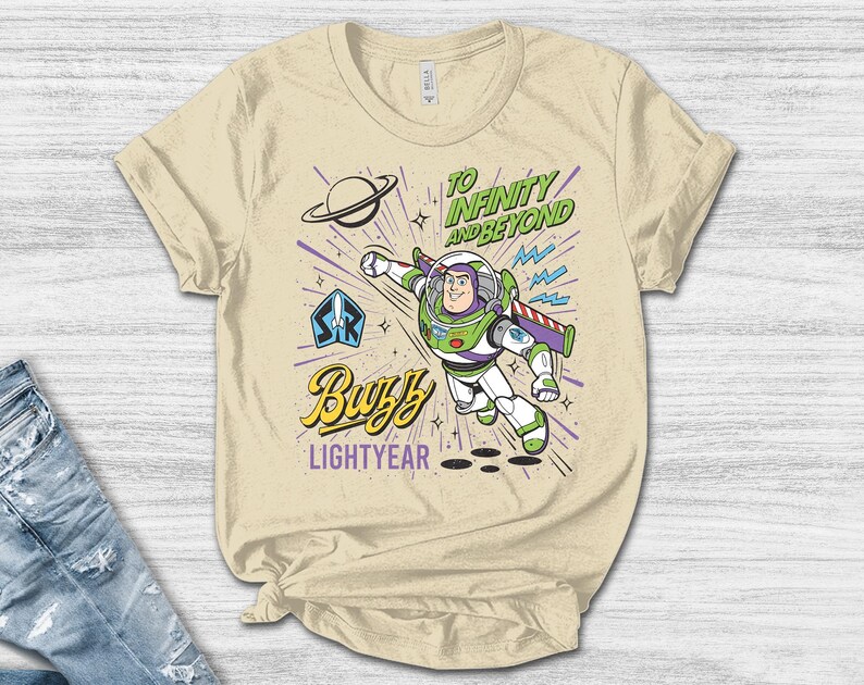 May include: A cream-colored t-shirt featuring a graphic of Buzz Lightyear in a flying pose. The design includes the text "TO INFINITY AND BEYOND" in green, "Buzz" in yellow script, and "LIGHTYEAR" in purple, along with a planet and star icons.