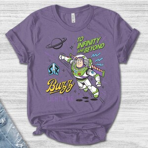 May include: A purple short-sleeve t-shirt featuring a graphic of Buzz Lightyear in a flying pose. The design includes the text "TO INFINITY AND BEYOND" in green, "Buzz" in yellow, and "LIGHTYEAR" in purple, along with a planet and star icons.