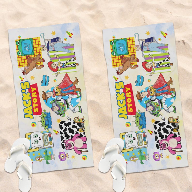 May include: Two white beach towels with a colorful cartoon design featuring characters and the text "JACK'S STORY". The towels also have white flip-flops at the bottom. The towels are on a sandy surface.