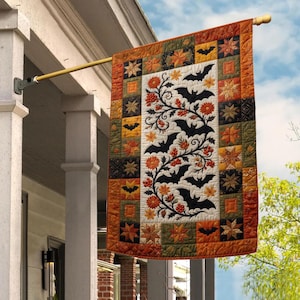 May include: A rectangular quilted garden flag hanging from a gold metal pole. The center panel features black bats and orange flowers on a white background. The border consists of a patchwork pattern of orange, green, and black squares.