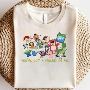 May include: A cream-colored t-shirt featuring a colorful graphic of various animated characters from the Toy Story franchise, including Woody, Buzz Lightyear, and Jessie. Below the characters, the text reads, "YOU'VE GOT A FRIEND IN ME."