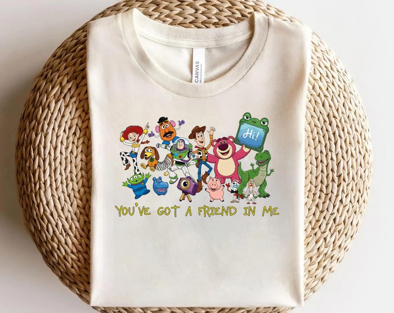 May include: A cream-colored t-shirt featuring a colorful graphic of various animated characters from the Toy Story franchise, including Woody, Buzz Lightyear, and Jessie. Below the characters, the text reads, "YOU'VE GOT A FRIEND IN ME."