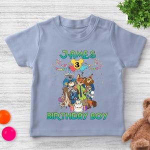 May include: A light blue short-sleeve t-shirt featuring a colorful graphic of Zootopia characters at a birthday party. The text reads "JAMES" at the top and "BIRTHDAY BOY" at the bottom, with a yellow number "3" inside a mouse-ear shape.