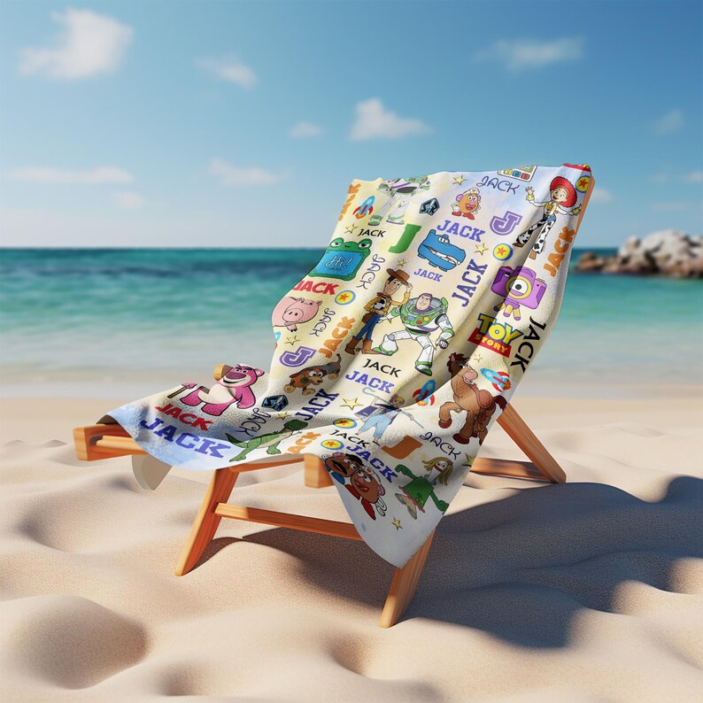 May include: A beach chair with a white towel featuring Toy Story characters and the name "JACK" in various fonts. The towel is draped over the wooden chair on a sandy beach with the ocean in the background.