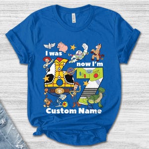 May include: Blue t-shirt featuring a colorful graphic with characters and the number 7 from the animated movie. The text reads "I was now I'm" and "Custom Name".