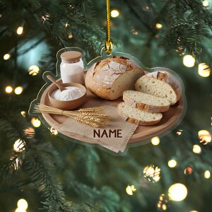 May include: A clear acrylic ornament featuring a bread-themed design. It includes a loaf of bread, sliced bread, a bowl of flour, a jar of flour, and wheat stalks. The word "NAME" is printed on a burlap-like fabric.