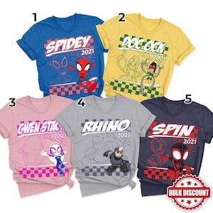 May include: Five short-sleeved t-shirts with superhero-themed graphics. The shirts are blue, yellow, pink, gray, and navy. Each shirt features a different character name and the year 2021. A red and white bulk discount sticker is in the bottom right corner.