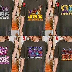 May include: Dark gray t-shirts with various cartoon character designs and Greek letter combinations. The shirts feature text like "HSS", "JOX", "OOZMA KAPPA", "PNK", "ΣMIC", and "ROR". Each design includes colorful illustrations.