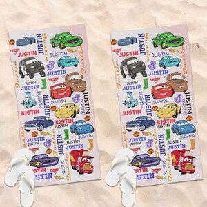 May include: Two rectangular beach towels with a Cars theme, featuring cartoon car illustrations and the name "Justin". White flip-flops are placed at the bottom of each towel. The towels are on a sandy surface. The towels are on a sandy surface.