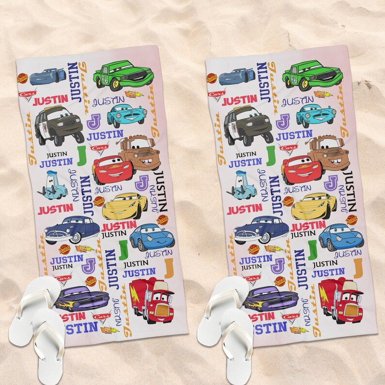 May include: Two rectangular beach towels with a Cars theme, featuring cartoon car illustrations and the name "Justin". White flip-flops are placed at the bottom of each towel. The towels are on a sandy surface. The towels are on a sandy surface.