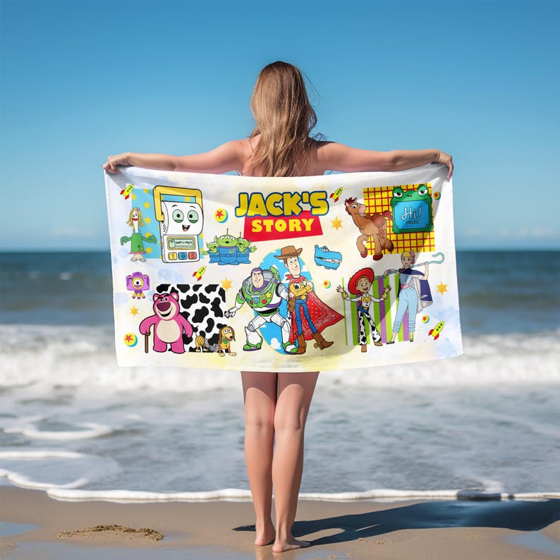 May include: A white beach towel featuring various cartoon characters and the text "JACK'S STORY". The towel has a colorful design with images of a robot, aliens, a bear, a dog, and other characters. The towel is held up by a person on a beach.