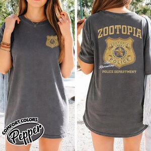 Puede incluir: Camiseta gris oscuro con un dise&ntilde;o de insignia de polic&iacute;a en la parte delantera y trasera. La insignia delantera dice "Police" y la trasera "Zootopia Police Department". La camiseta es de la marca Comfort Colors en color Pepper.