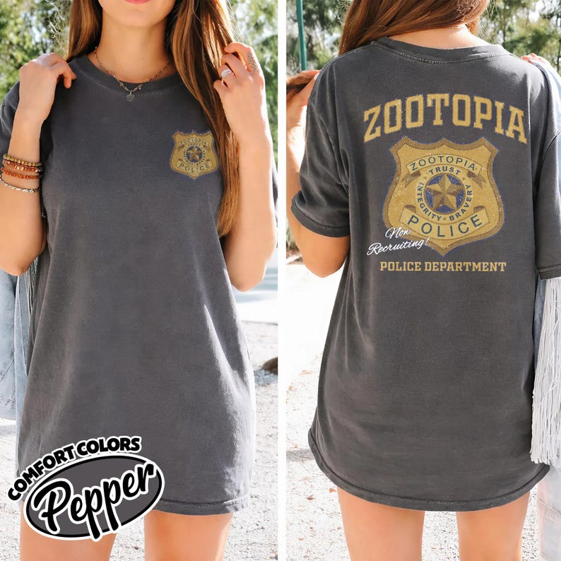 Puede incluir: Camiseta gris oscuro con un dise&ntilde;o de insignia de polic&iacute;a en la parte delantera y trasera. La insignia delantera dice "Police" y la trasera "Zootopia Police Department". La camiseta es de la marca Comfort Colors en color Pepper.