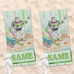 May include: Two rectangular beach towels featuring a cartoon space ranger character in a white, green, and purple suit. The design includes a sandcastle, a beach ball, a blue bucket, and the word NAME in green letters, paired with white flip-flops.