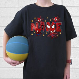 Puede incluir: Camiseta negra con la palabra "NAME" en rojo y blanco, con tem&aacute;tica de Spider-Man. Las letras est&aacute;n decoradas con telara&ntilde;as y peque&ntilde;as figuras de Spider-Man. Estrellas amarillas salpican las letras.
