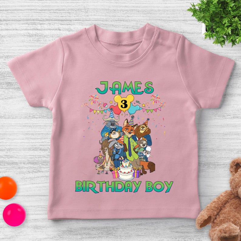 May include: A light pink t-shirt featuring a graphic print of Zootopia characters, a birthday cake, and colorful bunting. The text reads "JAMES" at the top, "3" inside a yellow mouse-ear shape, and "BIRTHDAY BOY" at the bottom in green letters.