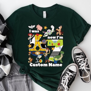 May include: Dark green t-shirt featuring characters from the animated film Toy Story. The shirt has the text "I was now I'm" and "Custom Name" with a large number seven. Black and white sneakers are at the bottom.