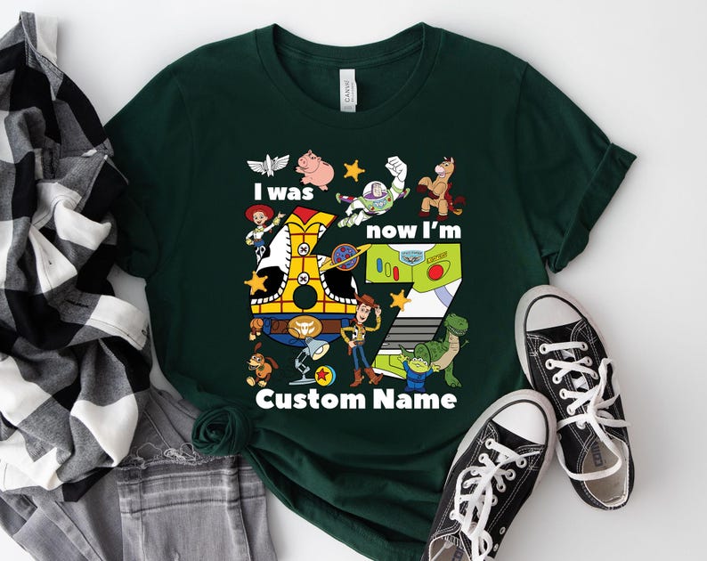May include: Dark green t-shirt featuring characters from the animated film Toy Story. The shirt has the text "I was now I'm" and "Custom Name" with a large number seven. Black and white sneakers are at the bottom.
