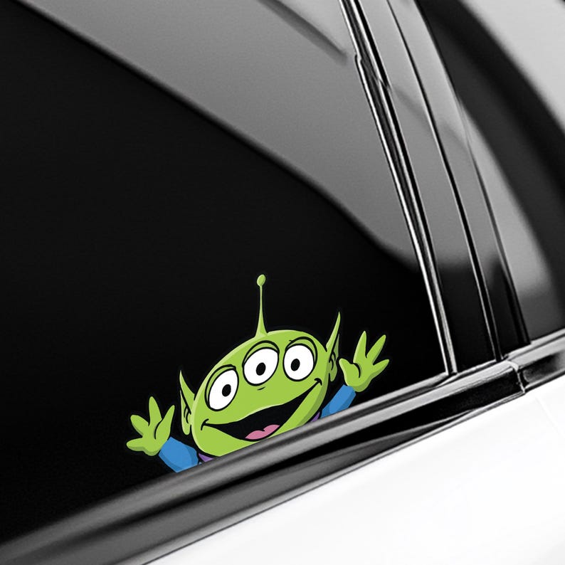 May include: A green alien character sticker with three eyes, a smiling mouth, and small hands. The alien is wearing a blue shirt and is positioned as if peeking out from behind a black surface, likely a car window.