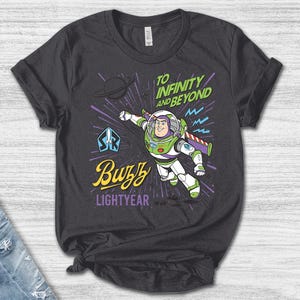 May include: A dark gray t-shirt featuring a graphic of Buzz Lightyear in a flying pose. The text reads "TO INFINITY AND BEYOND" in green, "Buzz" in yellow script, and "LIGHTYEAR" in purple. The design includes a planet, a space ranger logo, and purple speed lines.