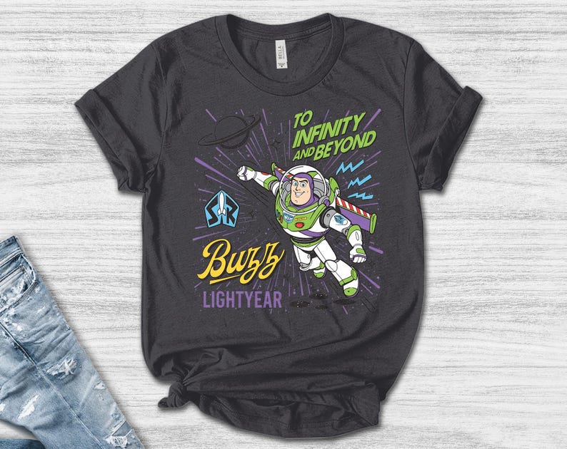 May include: A dark gray t-shirt featuring a graphic of Buzz Lightyear in a flying pose. The text reads "TO INFINITY AND BEYOND" in green, "Buzz" in yellow script, and "LIGHTYEAR" in purple. The design includes a planet, a space ranger logo, and purple speed lines.