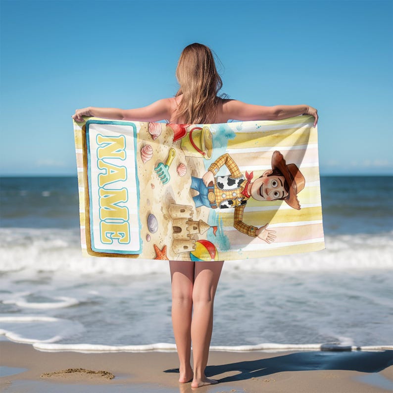 May include: A rectangular beach towel featuring a yellow and white striped background with a cartoon cowboy character, a sandcastle, seashells, a bucket, and a shovel. The towel includes a blue-bordered white rectangular box with the text NAME.