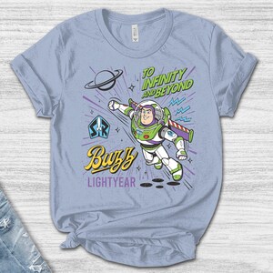 May include: A light blue t-shirt featuring a graphic of Buzz Lightyear in a flying pose. The design includes the text "TO INFINITY AND BEYOND", "Buzz", and "LIGHTYEAR" in green, yellow, and purple, along with a planet icon and a space ranger emblem.