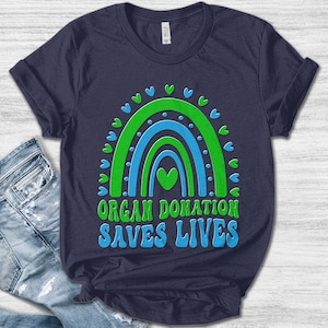 May include: Navy blue t-shirt featuring a green and blue rainbow design with heart accents. The text "ORGAN DONATION SAVES LIVES" is printed below the rainbow. The shirt is made of a soft material.