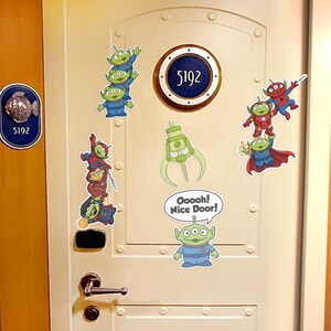 May include: A light beige door decorated with cartoon alien stickers. The stickers include green aliens in blue uniforms, a green claw, and aliens dressed as superheroes. The door has a circular plaque with the number 5192.