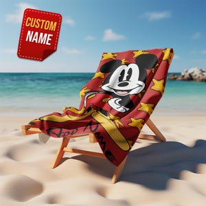 May include: A red beach towel draped over a wooden beach chair, featuring a large graphic of Mickey Mouse surrounded by yellow stars. The towel includes the text "ADD NAME" in a black script font, and a red tag displays "CUSTOM NAME".