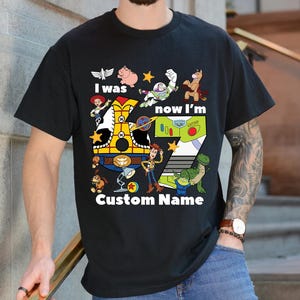 May include: Black t-shirt featuring a colorful graphic of characters from the Toy Story movie. The design includes the text "I was, now I'm" and "Custom Name" with the number 7. The shirt is a crew neck and is being worn.
