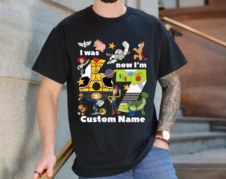 May include: Black t-shirt featuring a colorful graphic of characters from the Toy Story movie. The design includes the text "I was, now I'm" and "Custom Name" with the number 7. The shirt is a crew neck and is being worn.