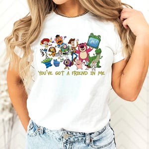 May include: A white short-sleeved t-shirt featuring a colorful graphic print of various animated characters from the Toy Story franchise. Below the characters, the text "YOU'VE GOT A FRIEND IN ME" is printed in a yellow, stylized font.