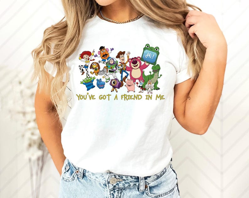 May include: A white short-sleeved t-shirt featuring a colorful graphic print of various animated characters from the Toy Story franchise. Below the characters, the text "YOU'VE GOT A FRIEND IN ME" is printed in a yellow, stylized font.