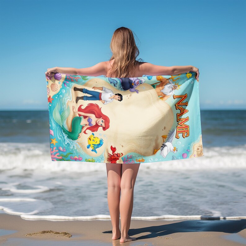 May include: A rectangular beach towel featuring a colorful illustration of characters from The Little Mermaid, including a mermaid, a prince, a crab, a fish, and a seagull on a sandy beach. The word NAME is printed in large orange letters.