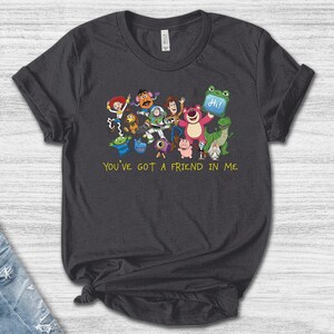 May include: A dark gray short-sleeve t-shirt featuring a colorful graphic of various animated toy characters and the yellow text "YOU'VE GOT A FRIEND IN ME" printed below the group. The shirt has a crew neckline and a standard fit.