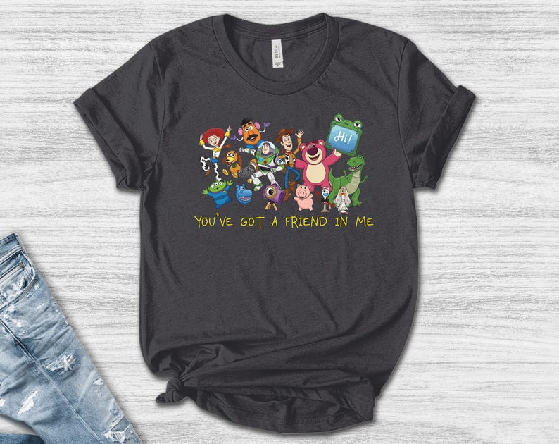 May include: A dark gray short-sleeve t-shirt featuring a colorful graphic of various animated toy characters and the yellow text "YOU'VE GOT A FRIEND IN ME" printed below the group. The shirt has a crew neckline and a standard fit.