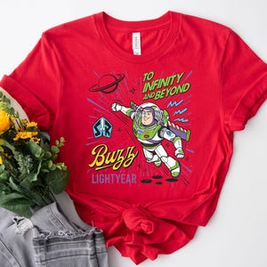 May include: A bright red short-sleeved t-shirt featuring a graphic of Buzz Lightyear in flight. The design includes the text TO INFINITY AND BEYOND, Buzz, and LIGHTYEAR, along with a planet, stars, and a space ranger emblem in blue, green, and yellow.